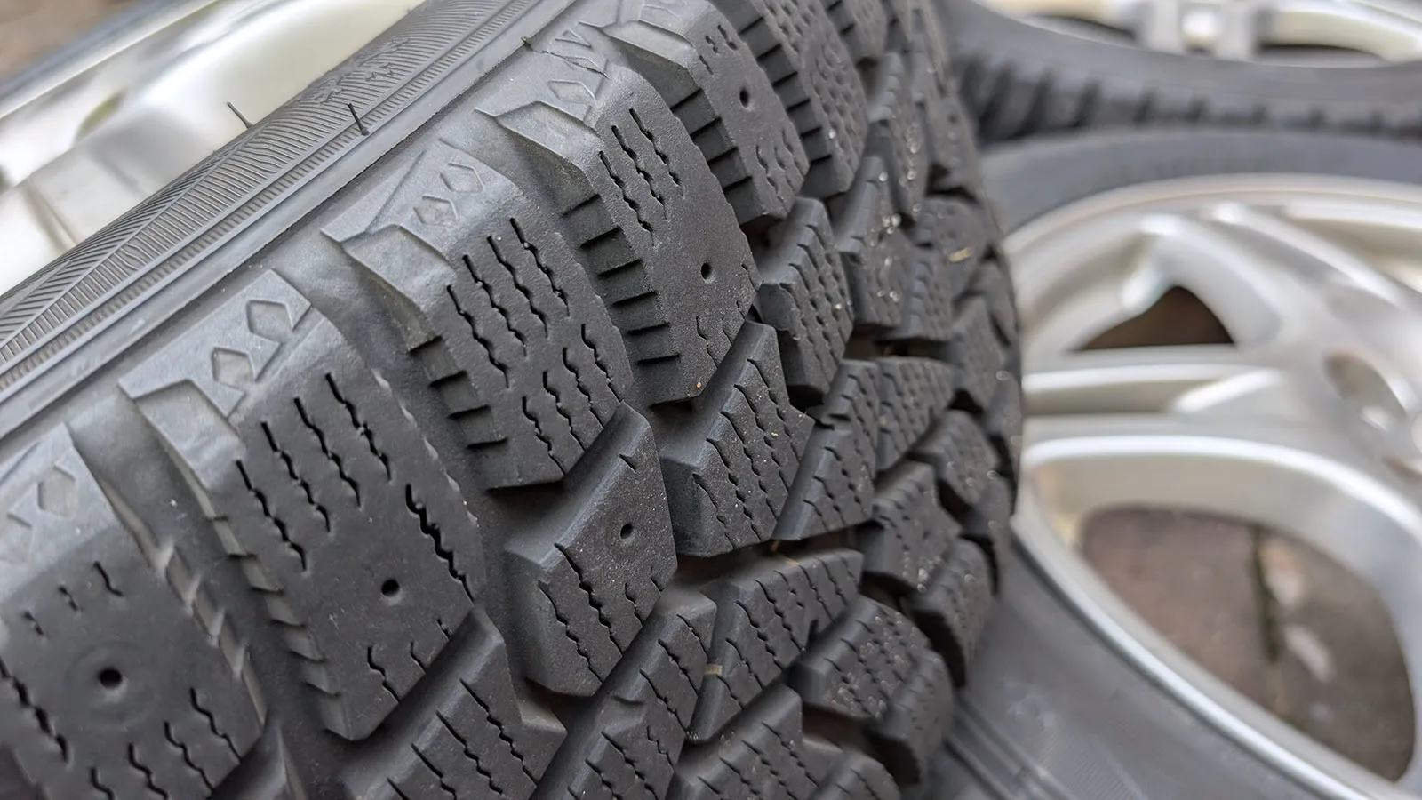 Winter Storage vs. Winter Tires: Which Makes Sense for a Second Car? 2 Close-up of winter tire tread showing dense siping, beside a stack of alloy wheels